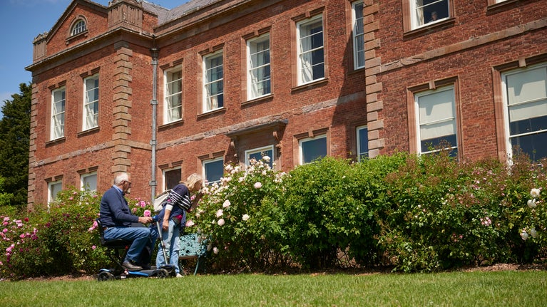 Two people admiring roses in bloom in front of a historic house made of red brick